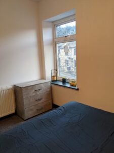 Property for sale by Auction A bedroom with a blue bedspread on the bed. A wooden chest of drawers is beside a large window. Items on the windowsill include a plant, candle holder, and decorative mirror. The window shows a view of buildings outside. Presented by Palace Auctions