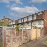 Property for sale by Auction A three-story brick building with wooden fences in the foreground. The sky is partly cloudy, and a modern apartment building is visible in the background. A small tree and some greenery are near the fence. Presented by Palace Auctions