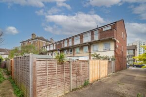 Property for sale by Auction A three-story brick building with wooden fences in the foreground. The sky is partly cloudy, and a modern apartment building is visible in the background. A small tree and some greenery are near the fence. Presented by Palace Auctions