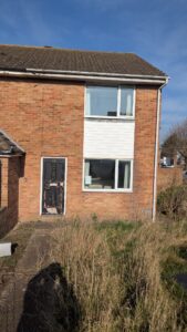 Property for sale by Auction A brick two-story house with a damaged front door, a white panel beneath a large window, and an overgrown front yard. The roof is tiled, and the sky is clear and sunny. Presented by Palace Auctions