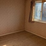 Property for sale by Auction An empty room with floral-patterned wallpaper and a wooden floor. There's a window with curtains on the right wall, letting in sunlight. A ceiling fan is partially visible at the top. Presented by Palace Auctions