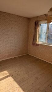 Property for sale by Auction An empty room with floral-patterned wallpaper and a wooden floor. There's a window with curtains on the right wall, letting in sunlight. A ceiling fan is partially visible at the top. Presented by Palace Auctions