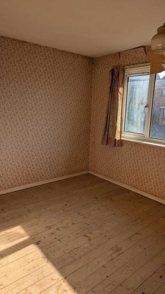 Property for sale by Auction An empty room with floral-patterned wallpaper and a wooden floor. There's a window with curtains on the right wall, letting in sunlight. A ceiling fan is partially visible at the top. Presented by Palace Auctions
