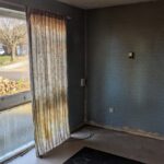 Property for sale by Auction Empty room with faded blue wallpaper and a dirty, partially open curtain next to a large window. The window offers a view of parked cars and bare trees on a residential street. The wall shows signs of dampness and peeling paint near the floor. Presented by Palace Auctions
