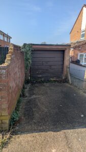 Property for sale by Auction A narrow driveway leads to a closed brown wooden garage door, flanked by brick walls. The left wall is partially damaged, with greenery growing on it. A chain-link fence adjoins the right side. Sunny day with a clear blue sky. Presented by Palace Auctions