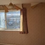 Property for sale by Auction A room with floral wallpaper is shown. The wallpaper is peeling at the top. There is a large window with pink curtains, and a white pendant light fixture hangs from the ceiling. Sunlight illuminates the room, and houses are visible outside. Presented by Palace Auctions