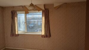 Property for sale by Auction A room with floral wallpaper is shown. The wallpaper is peeling at the top. There is a large window with pink curtains, and a white pendant light fixture hangs from the ceiling. Sunlight illuminates the room, and houses are visible outside. Presented by Palace Auctions