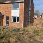 Property for sale by Auction A two-story brick house with a side window and a front door is surrounded by unkempt grass. The sky is clear and blue, and there's a paved area in the foreground with a shadow visible. Presented by Palace Auctions