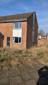 Property for sale by Auction A two-story brick house with a side window and a front door is surrounded by unkempt grass. The sky is clear and blue, and there's a paved area in the foreground with a shadow visible. Presented by Palace Auctions