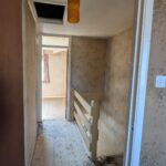 Property for sale by Auction A narrow, dimly lit hallway with patterned wallpaper and exposed floorboards. There is a small railing along the right side and an open doorway leading to a bright room with a window and patterned curtains. A yellow ceiling light hangs above. Presented by Palace Auctions