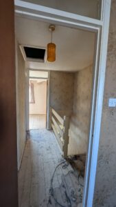 Property for sale by Auction A narrow, dimly lit hallway with patterned wallpaper and exposed floorboards. There is a small railing along the right side and an open doorway leading to a bright room with a window and patterned curtains. A yellow ceiling light hangs above. Presented by Palace Auctions