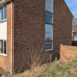 Property for sale by Auction A corner view of a two-story brick house with a sloped roof. The wall has vertical white siding sections. Wild grass and plants grow at the base. A clear blue sky is in the background. Presented by Palace Auctions