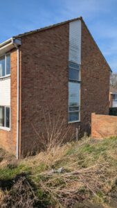 Property for sale by Auction A corner view of a two-story brick house with a sloped roof. The wall has vertical white siding sections. Wild grass and plants grow at the base. A clear blue sky is in the background. Presented by Palace Auctions
