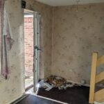 Property for sale by Auction Abandoned room with peeling wallpaper and litter on the floor. An open door partially shows a brick wall outside. A frayed curtain hangs beside the door. A wooden staircase is on the right. The space appears neglected and rundown. Presented by Palace Auctions