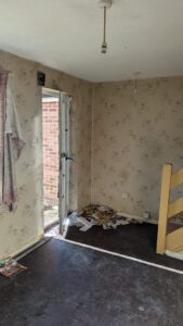 Property for sale by Auction Abandoned room with peeling wallpaper and litter on the floor. An open door partially shows a brick wall outside. A frayed curtain hangs beside the door. A wooden staircase is on the right. The space appears neglected and rundown. Presented by Palace Auctions