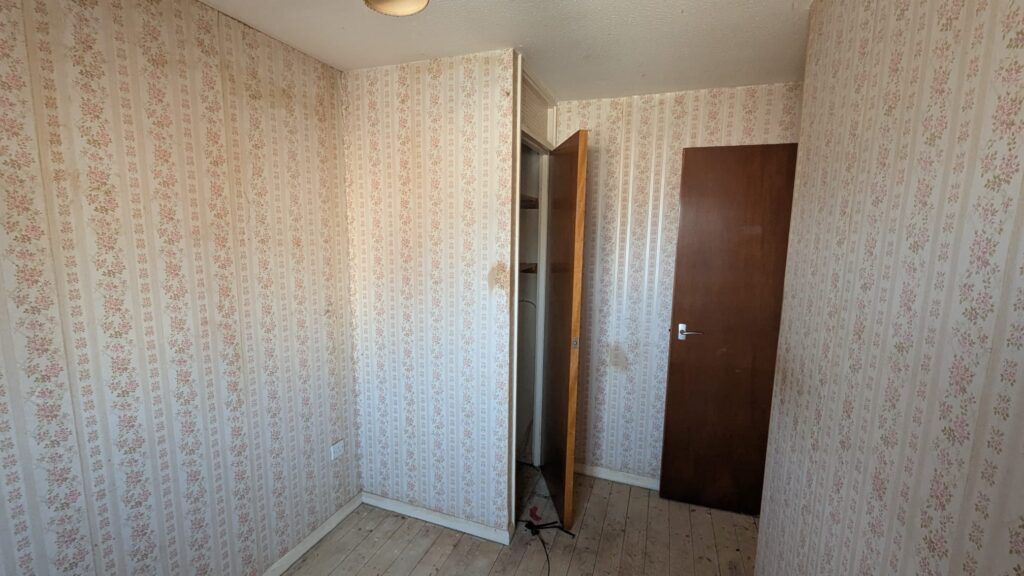 Property for sale by Auction A narrow, empty room with floral wallpaper and a light wooden floor. The room has two dark wooden doors, one slightly ajar revealing a small closet. A single ceiling light fixture is visible. Presented by Palace Auctions