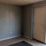 Property for sale by Auction An empty room with faded blue wallpaper, a dirty glass door, and a single window. The floor is bare, and a black rug lies in the corner. Sunlight filters through the window, illuminating the worn surfaces. Presented by Palace Auctions