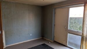 Property for sale by Auction An empty room with faded blue wallpaper, a dirty glass door, and a single window. The floor is bare, and a black rug lies in the corner. Sunlight filters through the window, illuminating the worn surfaces. Presented by Palace Auctions