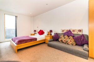 Property for sale by Auction A bedroom with a large window, featuring a wooden bed adorned with a red blanket and pillows. Two bedside tables with identical purple lamps flank the bed. A gray couch with floral and purple cushions is positioned nearby. The carpet is beige. Presented by Palace Auctions