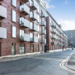 Property for sale by Auction Modern brick apartment building with multiple floors, featuring large rectangular windows and balconies. The street in front is empty, with clear skies above and another building visible in the background. Presented by Palace Auctions