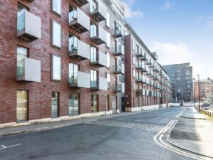 Property for sale by Auction Modern brick apartment building with multiple floors, featuring large rectangular windows and balconies. The street in front is empty, with clear skies above and another building visible in the background. Presented by Palace Auctions
