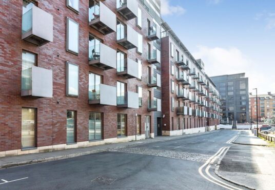 Property for sale by Auction Modern brick apartment building with multiple floors, featuring large rectangular windows and balconies. The street in front is empty, with clear skies above and another building visible in the background. Presented by Palace Auctions