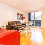 Property for sale by Auction Modern living room with red and black sofas, a glass coffee table, a flat-screen TV on a white stand, and a dining area with glass-top table and chairs. Large window offers cityscape view. Bright artwork decorates the wall. Presented by Palace Auctions