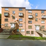 Property for sale A three-story brick apartment building with multiple windows, balconies, and satellite dishes—ideal for those seeking an investment studio flat. Two main entrances have ramps and stairs, with grass, a sidewalk in front, and trees to the left. Presented by Palace Auctions