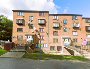 Property for sale A three-story brick apartment building with multiple windows, balconies, and satellite dishes—ideal for those seeking an investment studio flat. Two main entrances have ramps and stairs, with grass, a sidewalk in front, and trees to the left. Presented by Palace Auctions