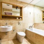 Property for sale by Auction A modern bathroom with beige tiles, a wall-mounted sink, and a toilet. There's a bathtub with a shower fixture, a wall mirror above the sink, and a towel rack on the left. A small trash bin is under the sink. Presented by Palace Auctions