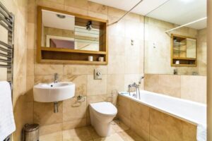 Property for sale by Auction A modern bathroom with beige tiles, a wall-mounted sink, and a toilet. There's a bathtub with a shower fixture, a wall mirror above the sink, and a towel rack on the left. A small trash bin is under the sink. Presented by Palace Auctions