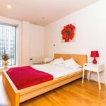 Property for sale by Auction A minimalist bedroom with a wooden floor and a platform bed featuring red and white bedding. A large red floral artwork hangs above the bed. A red lamp and small white tables flank the bed. A tall window with vertical blinds is on the left. Presented by Palace Auctions