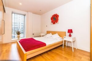 Property for sale by Auction A minimalist bedroom with a wooden floor and a platform bed featuring red and white bedding. A large red floral artwork hangs above the bed. A red lamp and small white tables flank the bed. A tall window with vertical blinds is on the left. Presented by Palace Auctions