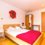 Property for sale by Auction A minimalist bedroom with a wooden bed, white bedding, and a red blanket. Two small white nightstands with red lamps flank the bed. A wardrobe and a desk are against the wall. A painting of a red flower hangs above the bed on a white wall. Presented by Palace Auctions
