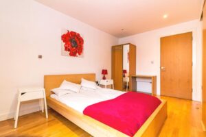 Property for sale by Auction A minimalist bedroom with a wooden bed, white bedding, and a red blanket. Two small white nightstands with red lamps flank the bed. A wardrobe and a desk are against the wall. A painting of a red flower hangs above the bed on a white wall. Presented by Palace Auctions