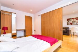 Property for sale by Auction Bedroom with a wooden bed, white bedding, and a red blanket. A red lamp is on the bedside table. There's a wardrobe with sliding doors and a mirror. An open door reveals a living area with a sofa, table, and floral artwork. Presented by Palace Auctions