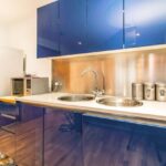 Property for sale by Auction A modern kitchen with glossy blue cabinets, a double sink, and a stainless steel backsplash. On the counter are canisters and a microwave. A white refrigerator stands beside a wooden shelf unit on the left. The floor is light-colored wood. Presented by Palace Auctions