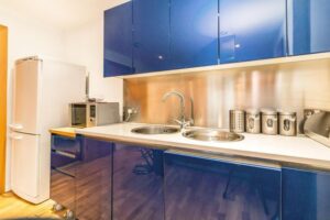 Property for sale by Auction A modern kitchen with glossy blue cabinets, a double sink, and a stainless steel backsplash. On the counter are canisters and a microwave. A white refrigerator stands beside a wooden shelf unit on the left. The floor is light-colored wood. Presented by Palace Auctions
