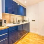 Property for sale by Auction Modern kitchen with glossy blue cabinets, a stainless steel backsplash, and wooden flooring. A microwave is placed on a small counter with drawers. A kettle and toaster sit on the countertop. A wall art piece hangs on a white wall. Presented by Palace Auctions