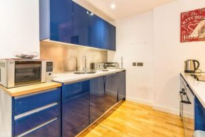 Property for sale by Auction Modern kitchen with glossy blue cabinets, a stainless steel backsplash, and wooden flooring. A microwave is placed on a small counter with drawers. A kettle and toaster sit on the countertop. A wall art piece hangs on a white wall. Presented by Palace Auctions