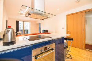 Property for sale by Auction Modern kitchen with a sleek electric stovetop on a blue island, featuring a glass range hood above. A stainless steel kettle sits on the counter. In the background, a bright living area with a red sofa and a window is visible. Presented by Palace Auctions