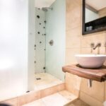 Property for sale by Auction Modern bathroom with a wood-framed mirror above a wooden countertop. A white vessel sink and wall-mounted faucet are on the right. The shower features frosted glass and beige tiles surround the area, creating a clean and minimalist look. Presented by Palace Auctions