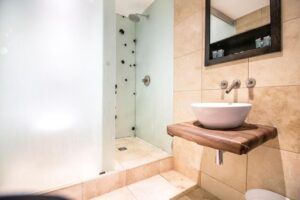 Property for sale by Auction Modern bathroom with a wood-framed mirror above a wooden countertop. A white vessel sink and wall-mounted faucet are on the right. The shower features frosted glass and beige tiles surround the area, creating a clean and minimalist look. Presented by Palace Auctions