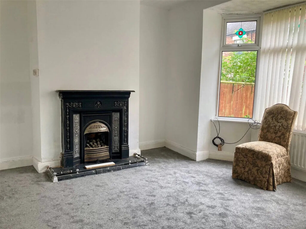 Property for sale A sparsely furnished room in a Stanley residential property with gray carpet, a black decorative fireplace, a single upholstered chair, and a window with vertical blinds showing greenery outside. A loose cable hangs near the window. Presented by Palace Auctions