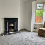 Property for sale A sparsely furnished room in a Stanley residential property with gray carpet, a black decorative fireplace, a single upholstered chair, and a window with vertical blinds showing greenery outside. A loose cable hangs near the window. Presented by Palace Auctions