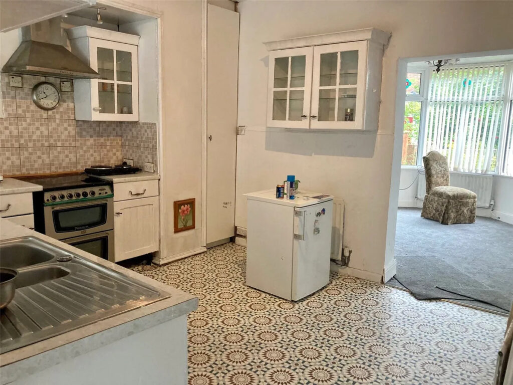 Property for sale A kitchen with patterned tile flooring, white cabinets, a small white fridge, stove, and a stainless steel sink. An adjacent room with carpet and a single patterned chair is visible—ideal for a Rose Avenue family home in County Durham. Presented by Palace Auctions