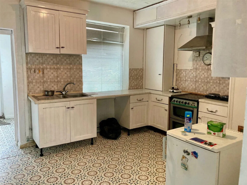 Property for sale A kitchen in a Stanley 3-bed terrace with white cabinets, a tile backsplash, gas stove and oven, stainless steel range hood, small fridge, patterned floor tiles, window blinds, and various items on the counters. Presented by Palace Auctions