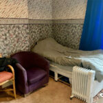Property for sale A cozy bedroom in a Stanley 3-bed terrace features a single bed with gray bedding, a purple armchair, wooden chair with jacket, portable white heater, blue curtain at the window, and patterned wallpaper. Presented by Palace Auctions