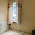 Property for sale A small, empty room with beige wallpaper, a single window with cream curtains, a white radiator, and some clutter on the windowsill. This Stanley property for sale appears to be in need of renovation. Presented by Palace Auctions