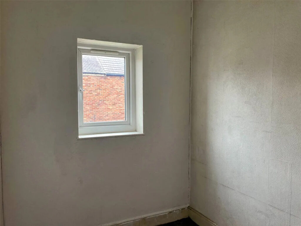 Property for sale A small, empty room with plain off-white walls and a single square window showing part of a brick building outside—typical of what you might find in a County Durham house auction. The room appears undecorated and dimly lit. Presented by Palace Auctions